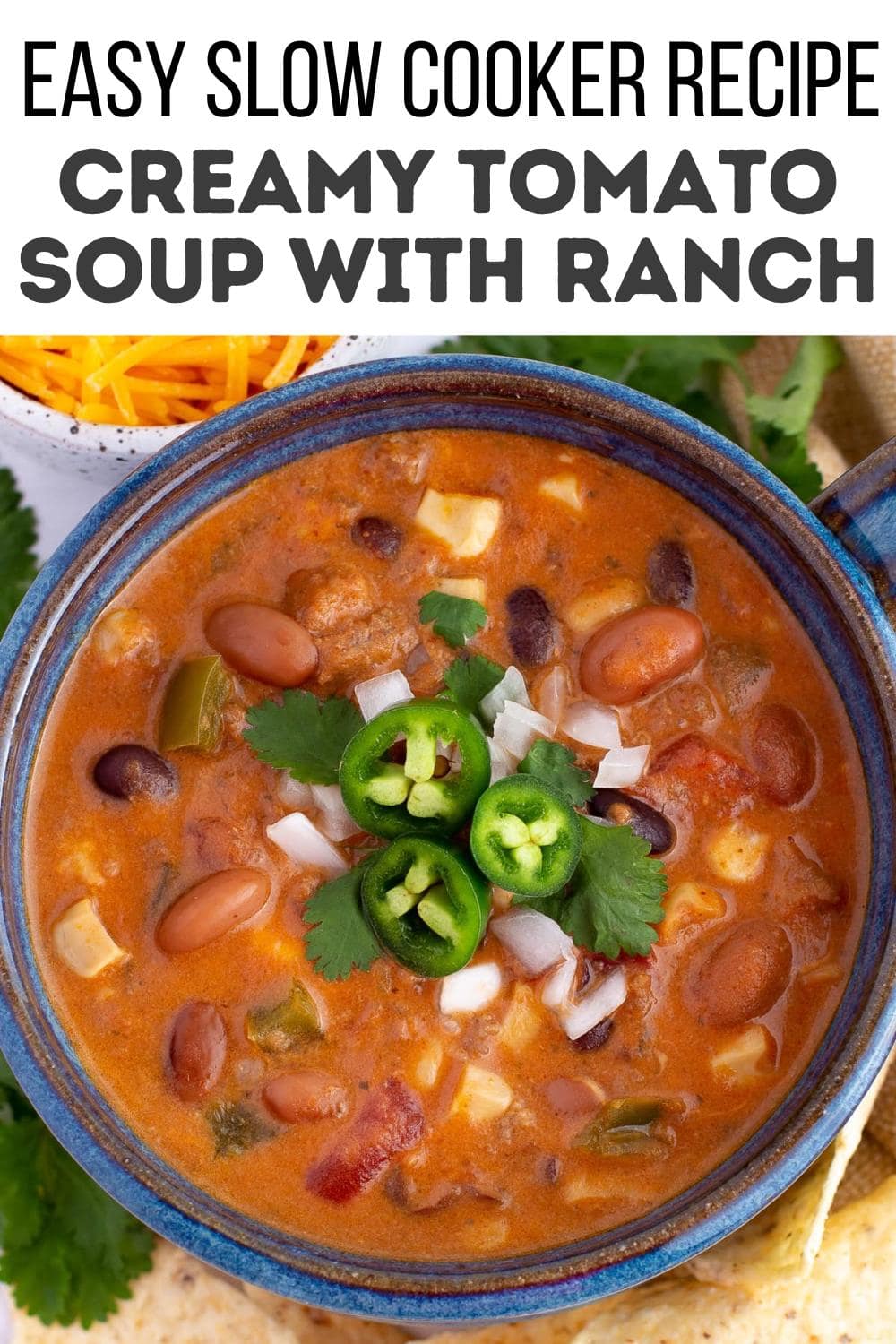 A bowl of creamy tomato soup with beans, topped with sliced jalapeños, chopped onions, and cilantro—including a hint of canned cowboy candy recipe inspiration—sits on a blue plate. Text above reads "Easy Slow Cooker Recipe: Creamy Tomato Soup with Ranch.