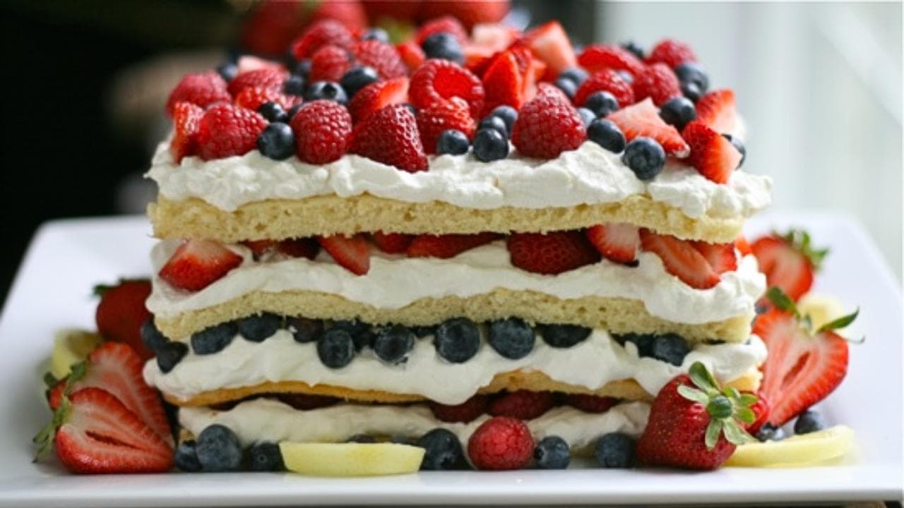 A three-layer sponge cake topped and filled with whipped cream and fresh berries including strawberries, blueberries, and raspberries, arranged on a white plate with extra fruit around the base.