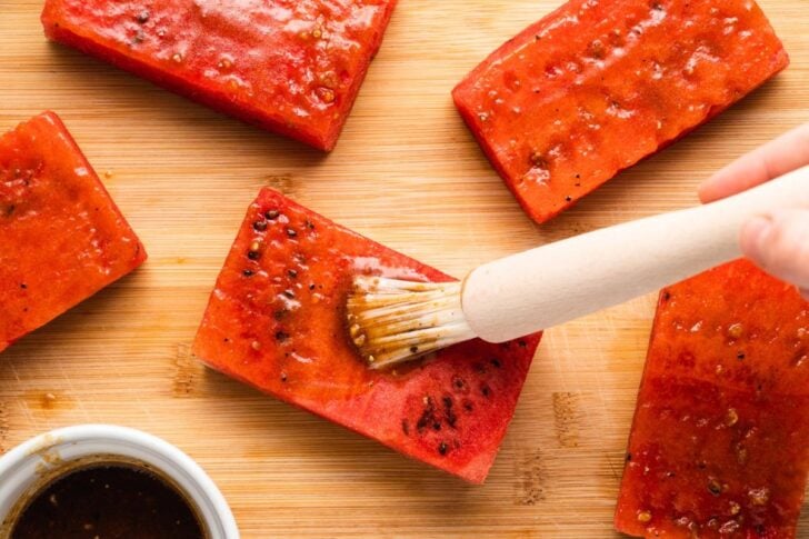 Rectangular slices of watermelon are being brushed with a brown marinade on a wooden cutting board. A bowl of the marinade is visible in the corner.
