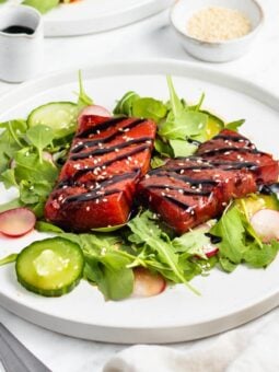 A white plate with mixed greens, sliced cucumber, and radishes, topped with two pieces of marinated watermelon, drizzled with dark sauce, and sprinkled with sesame seeds. A small pitcher and bowl are in the background.