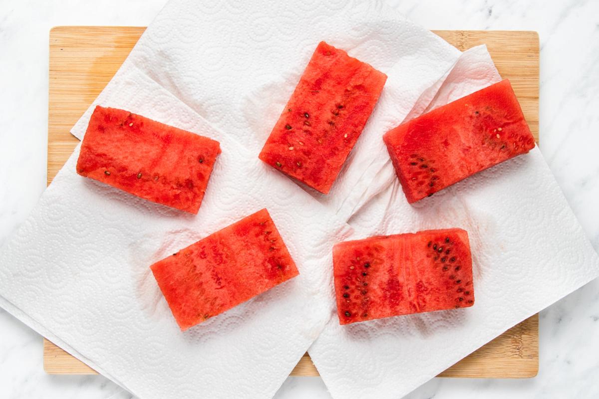 Five rectangular slices of watermelon placed on white paper towels, arranged on a wooden cutting board atop a white marble surface.
