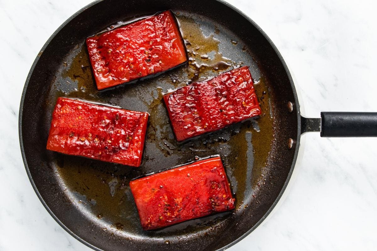 Four rectangular slices of glazed watermelon are searing in a black frying pan with some oil, on a white marble surface. The watermelon pieces are browned on the edges, resembling cooked fish fillets.