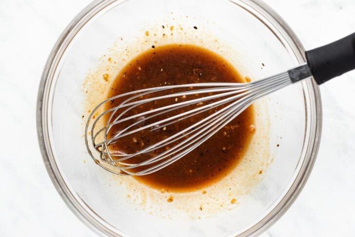 A metal whisk rests in a glass bowl containing a brown, seasoned liquid mixture with visible spices and small garlic pieces on a white surface.