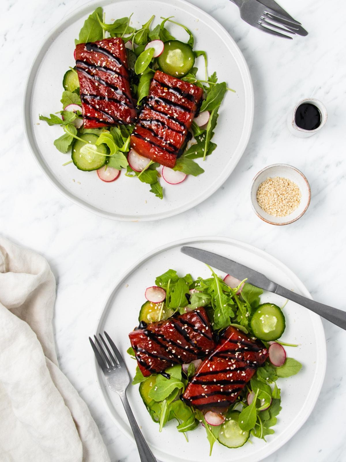 Two white plates with green salads topped with glazed tofu, sliced cucumbers, radishes, arugula, and sesame seeds. A small bowl of sesame seeds, a dish of dark sauce, and utensils are also visible on a white surface.