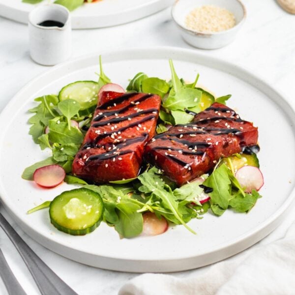 A white plate with mixed greens, sliced cucumber, and radishes, topped with two pieces of marinated watermelon, drizzled with dark sauce, and sprinkled with sesame seeds. A small pitcher and bowl are in the background.