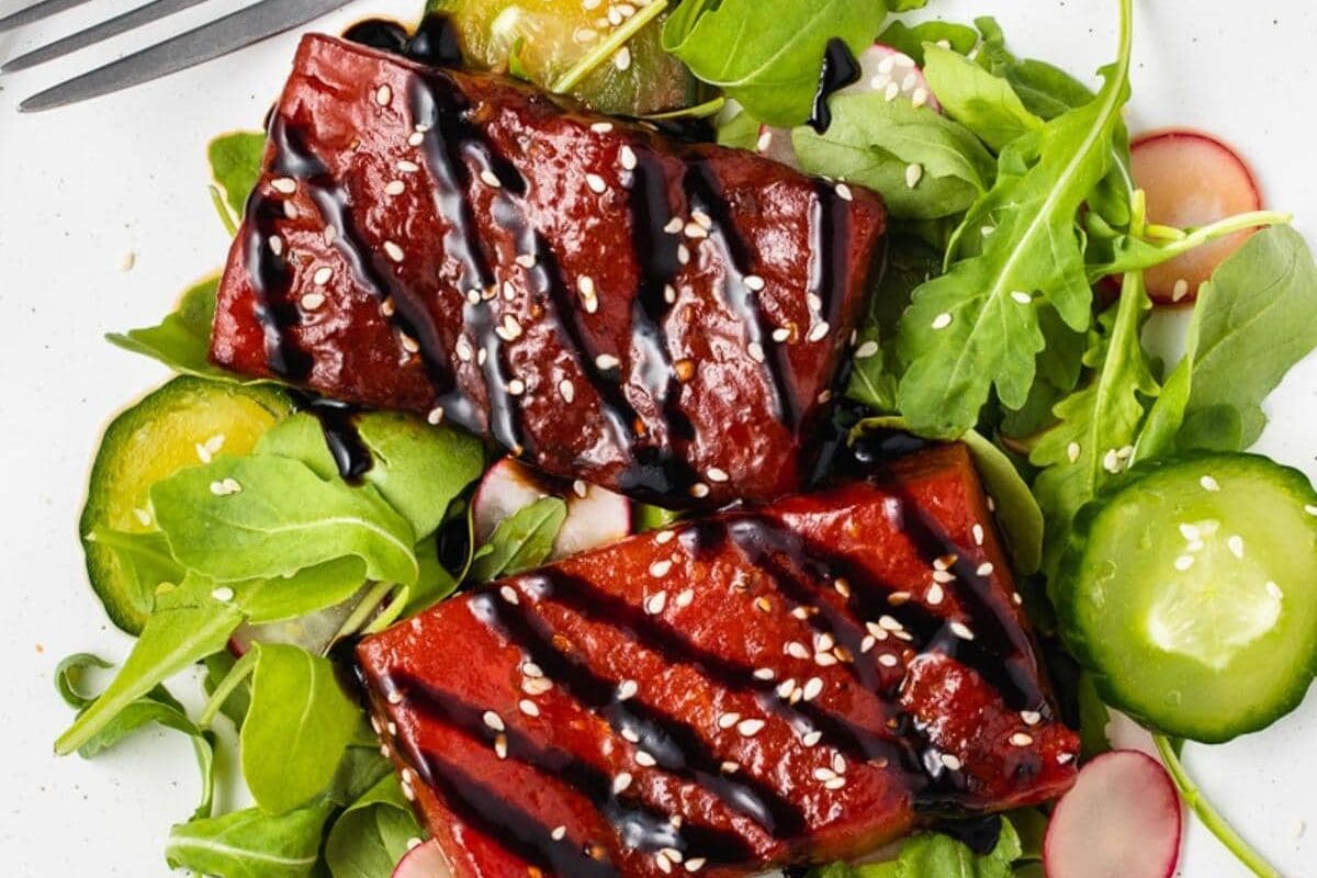 Two slices of glazed tofu on a bed of arugula, cucumber, and radish slices, drizzled with dark sauce and sprinkled with sesame seeds, served on a white plate with a fork.