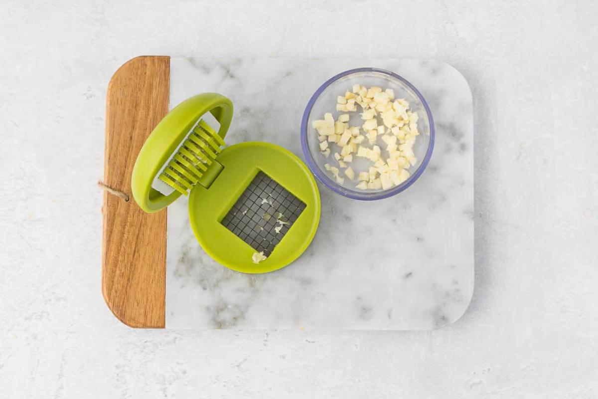 A green garlic dicer sits open on a marble and wood cutting board, with diced garlic pieces inside a small clear container nearby.