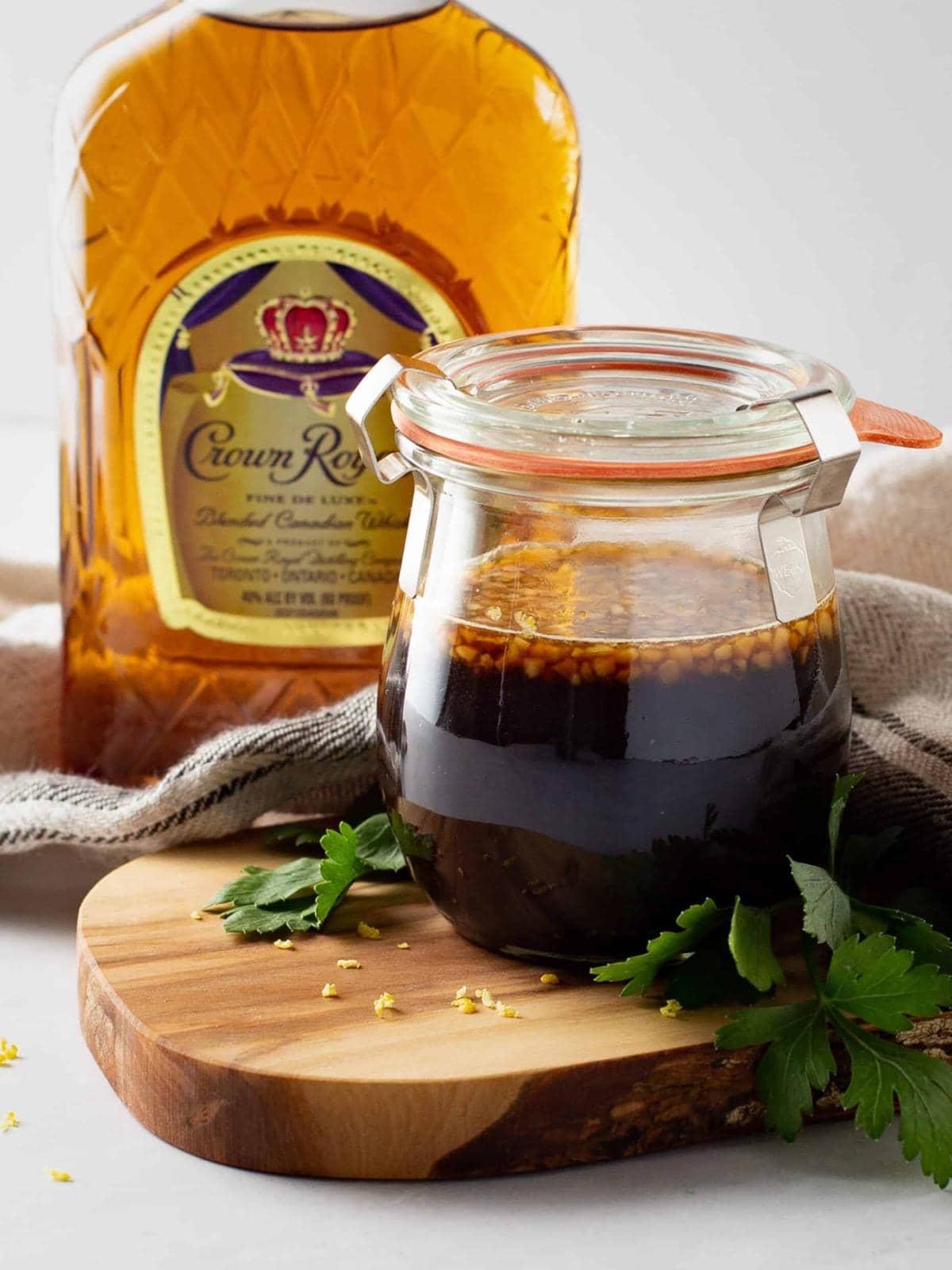 A glass jar filled with a dark marinade sits on a wooden board next to a bottle of Crown Royal whisky, with parsley leaves scattered nearby.