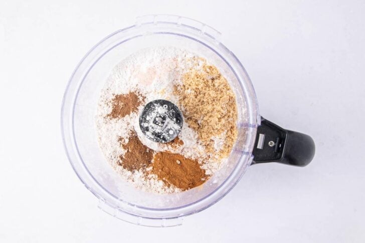 A food processor bowl filled with flour, ground cinnamon, ground ginger, and other dry ingredients, viewed from above on a white background.