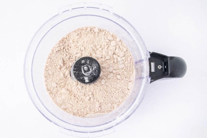 Top view of a food processor filled with finely ground flour or powder, with the blade attachment visible in the center, set against a white background.
