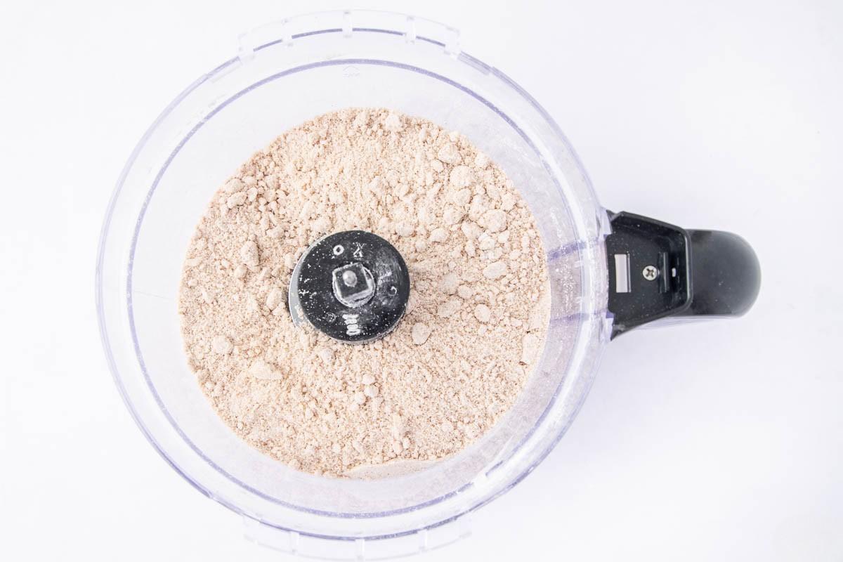 Top view of a food processor filled with finely ground flour or powder, with the blade attachment visible in the center, set against a white background.