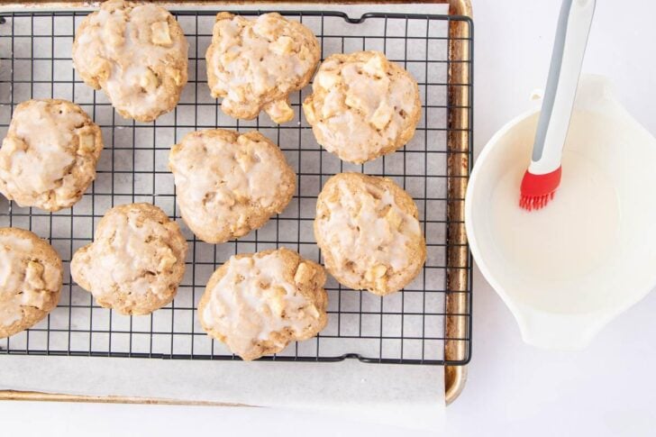Nine iced cookies cool on a wire rack set over a baking sheet. Beside them is a bowl of glaze with a red pastry brush resting inside. The background is bright and white.