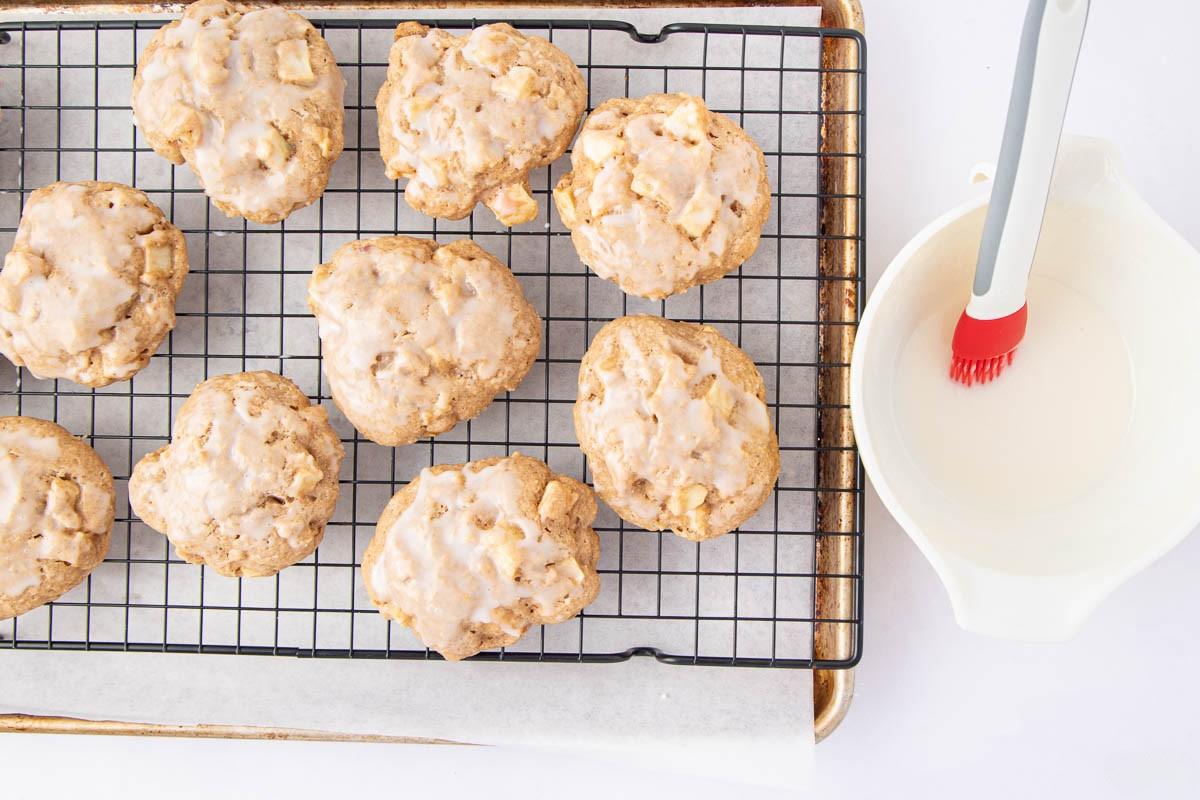 Nine iced cookies cool on a wire rack set over a baking sheet. Beside them is a bowl of glaze with a red pastry brush resting inside. The background is bright and white.