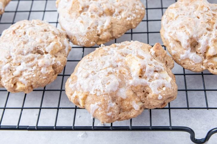Iced apple cookies with a light glaze cooling on a black wire rack over a white surface.