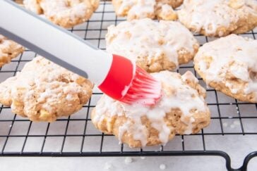 A red and white pastry brush is spreading white glaze onto round cookies cooling on a black wire rack. Other glazed cookies are visible in the background.