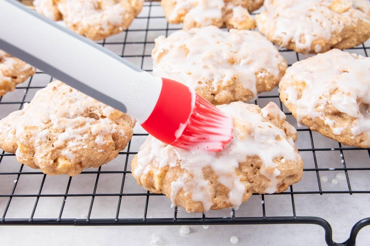 A red and white pastry brush is spreading white glaze onto round cookies cooling on a black wire rack. Other glazed cookies are visible in the background.