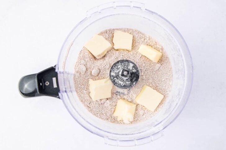 A food processor bowl containing a mixture of flour and spices with several chunks of butter placed on top, viewed from above.