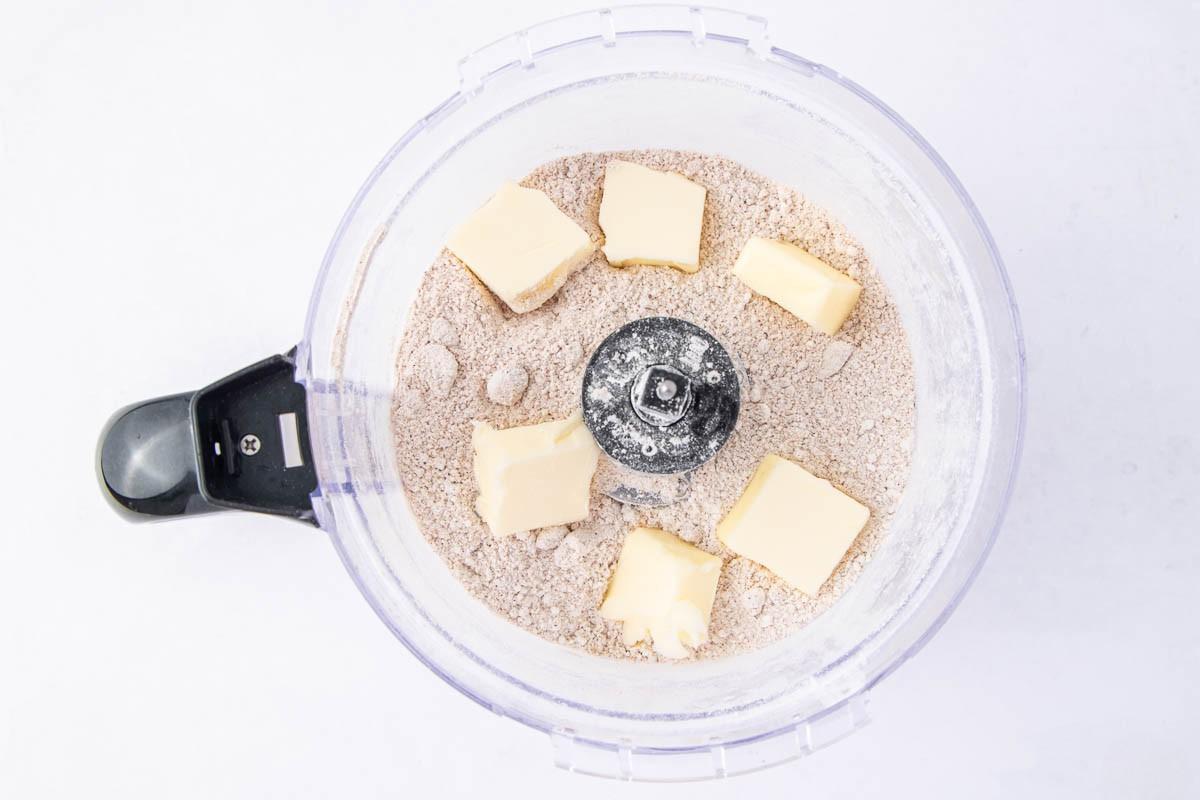 A food processor bowl containing a mixture of flour and spices with several chunks of butter placed on top, viewed from above.