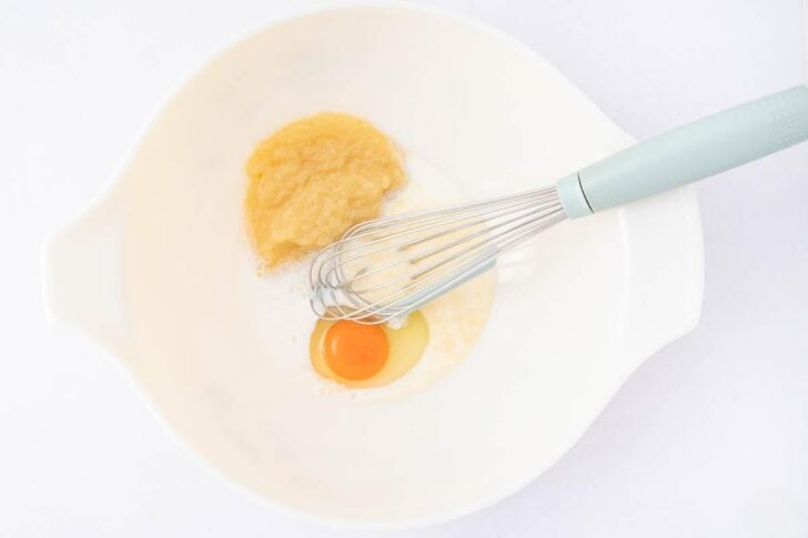 A white mixing bowl with a raw egg, applesauce, and milk inside. A metal whisk with a light blue handle rests in the bowl on a white background.