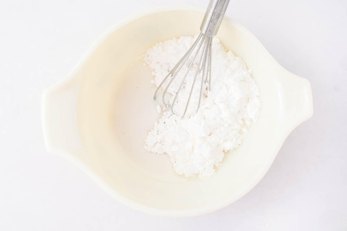 A metal whisk rests in a white mixing bowl containing a mound of white powder, likely flour or powdered sugar, on a light surface.