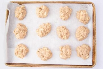 Twelve unbaked cookie dough mounds are evenly spaced on a parchment-lined baking sheet, ready to be baked. The dough appears chunky, possibly containing pieces of fruit or nuts.