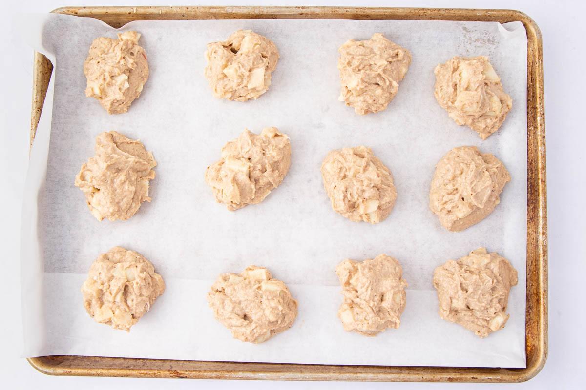 Twelve unbaked cookie dough mounds are evenly spaced on a parchment-lined baking sheet, ready to be baked. The dough appears chunky, possibly containing pieces of fruit or nuts.