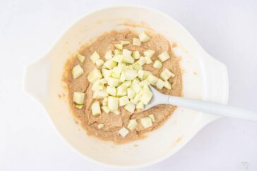 A white mixing bowl containing a light brown batter with chopped green apple pieces and a white spatula resting inside.