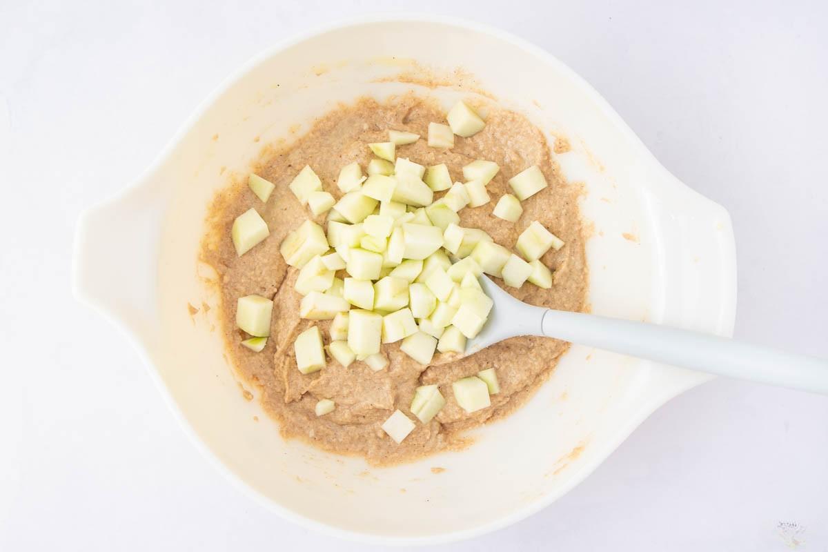 A white mixing bowl containing a light brown batter with chopped green apple pieces and a white spatula resting inside.