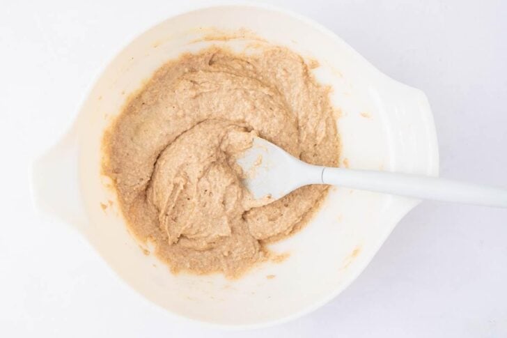 A white mixing bowl contains tan-colored batter being stirred with a white spatula on a white background.