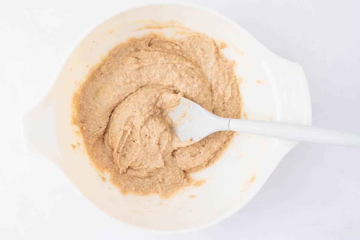 A white mixing bowl contains tan-colored batter being stirred with a white spatula on a white background.