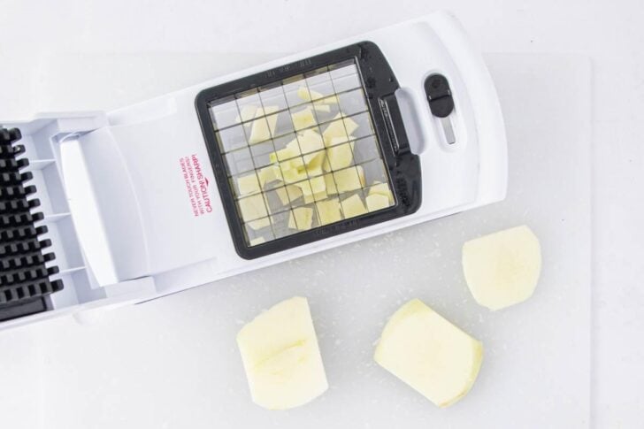 A white vegetable chopper with diced pieces of potato inside and three peeled potato chunks on a white cutting board next to it.