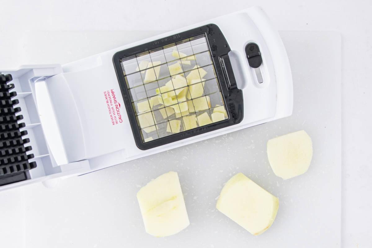 A white vegetable chopper with diced pieces of potato inside and three peeled potato chunks on a white cutting board next to it.