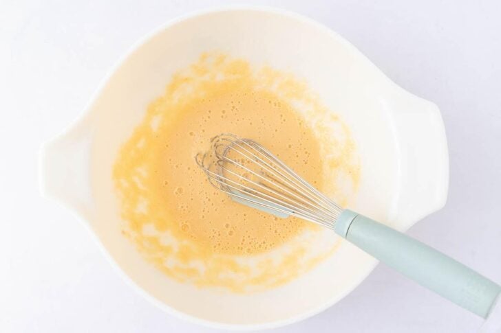 A white mixing bowl containing a whisk and a light, frothy yellow mixture, likely eggs being beaten, is placed on a white surface.