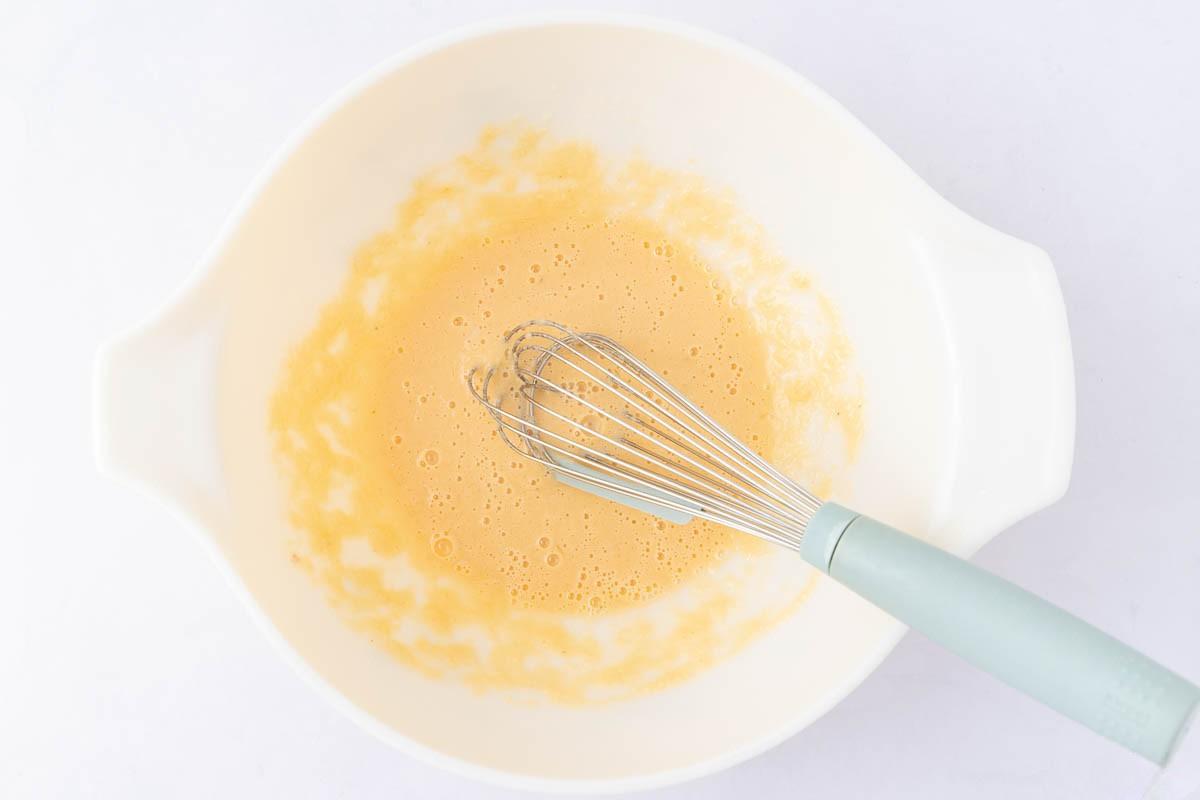 A white mixing bowl containing a whisk and a light, frothy yellow mixture, likely eggs being beaten, is placed on a white surface.