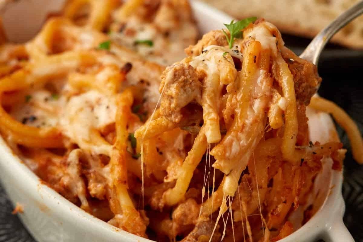 A close-up of baked spaghetti in a white dish, topped with melted cheese and herbs, with a fork lifting a cheesy portion.