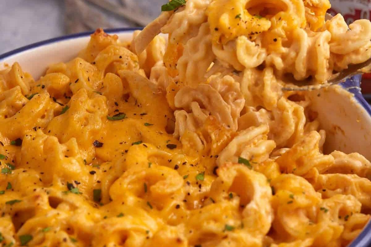 A close-up of creamy macaroni and cheese in a bowl, with a fork lifting a portion. The dish is topped with melted cheese, black pepper, and garnished with chopped herbs.
