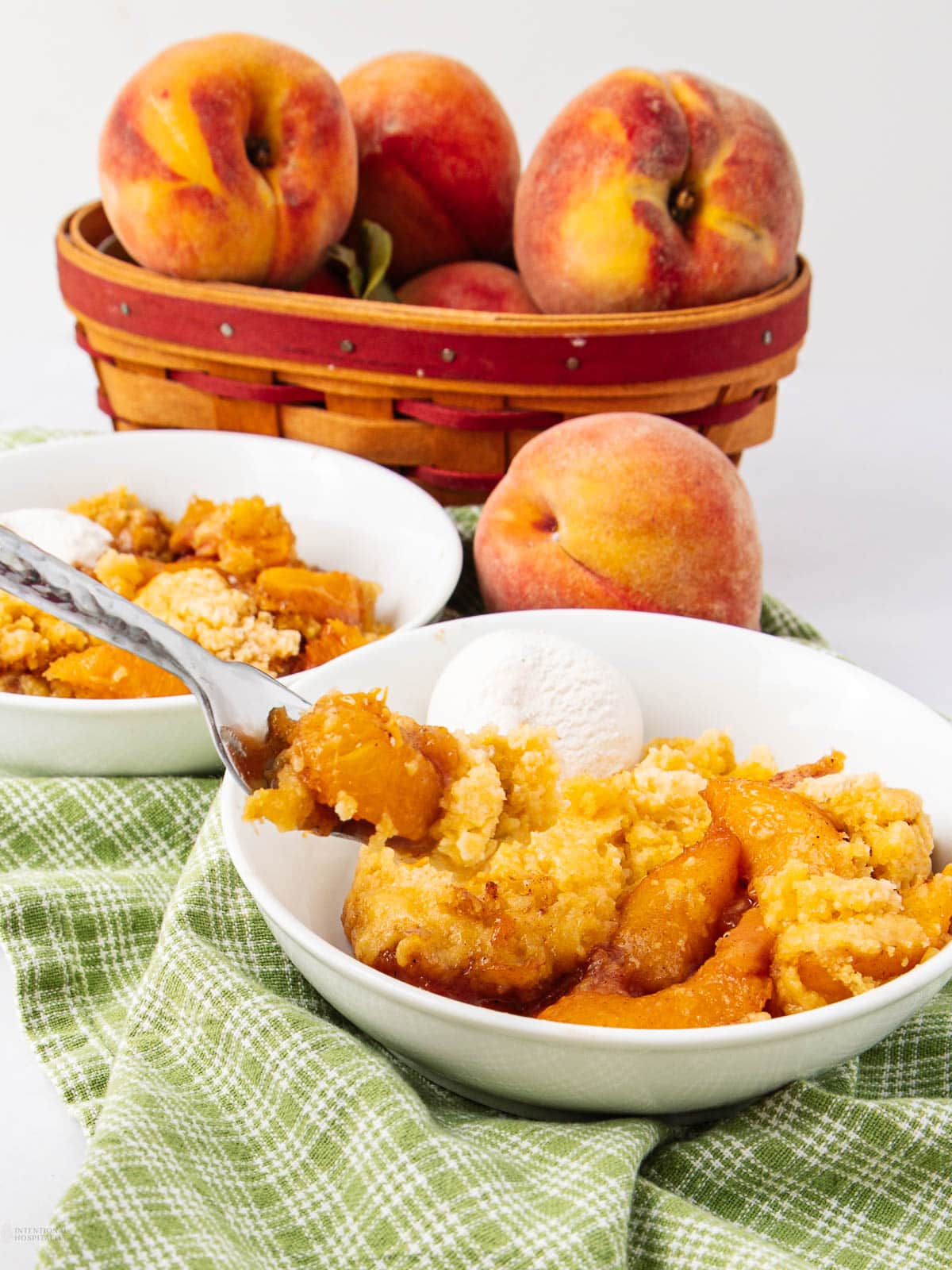 A bowl of peach cobbler with a scoop of vanilla ice cream sits on a green plaid cloth, with whole peaches and a basket of peaches in the background.