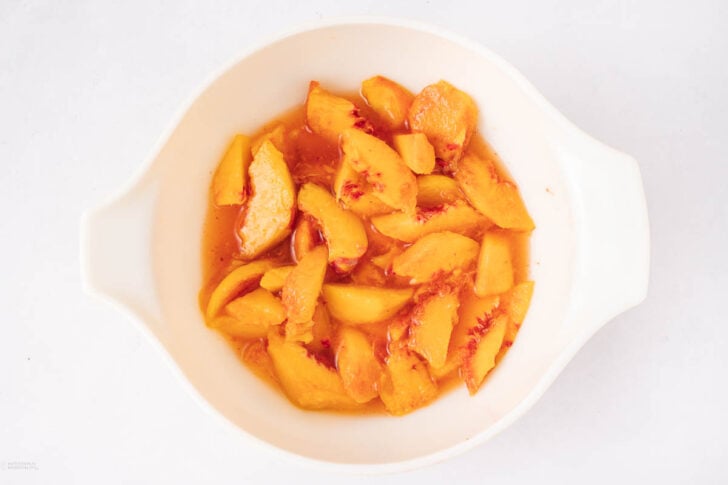 Sliced peaches with juice in a white bowl, viewed from above on a plain white background.