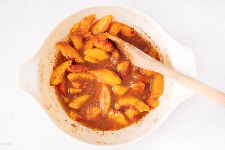 Sliced peaches mixed with brown sugar and spices in a white bowl, stirred with a wooden spoon, on a white background.