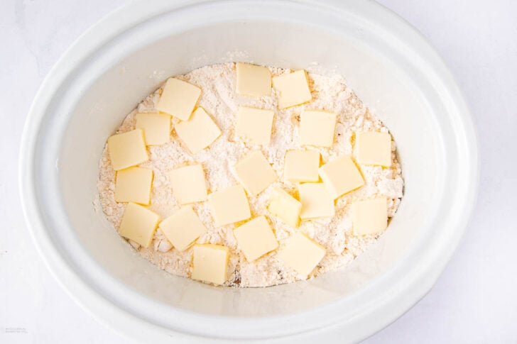 A white slow cooker filled with flour, topped with evenly spaced slices of butter.