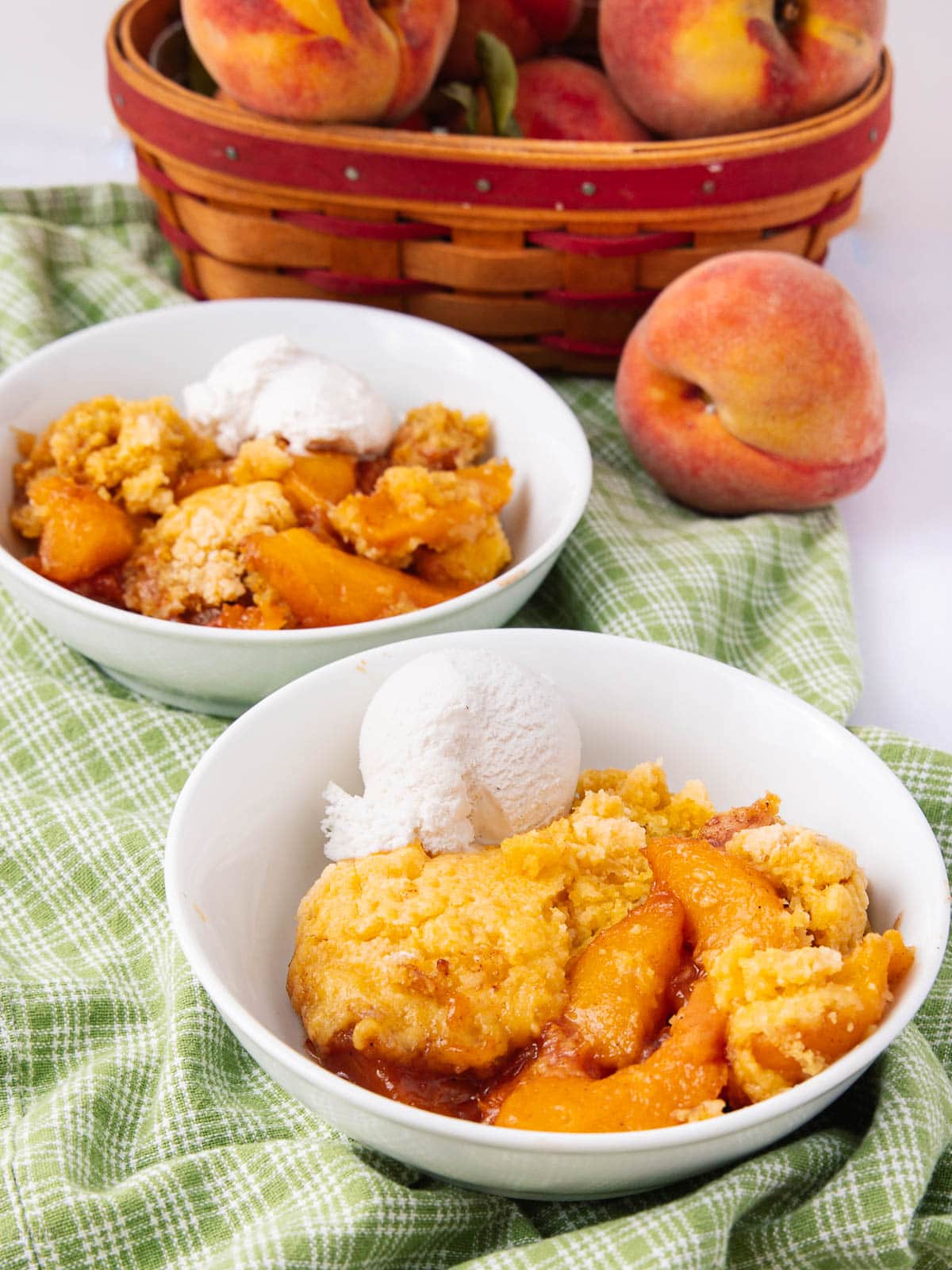 Two bowls of peach cobbler topped with vanilla ice cream sit on a green checkered cloth, with a basket of fresh peaches and a whole peach in the background.