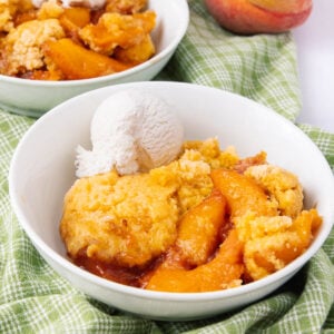 A bowl of peach cobbler with golden biscuit topping, served with a scoop of vanilla ice cream, sits on a green checkered cloth. Another bowl and a fresh peach are visible in the background.