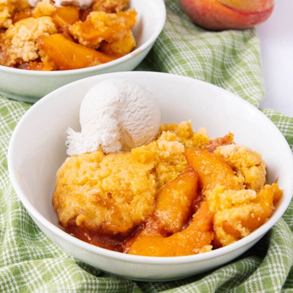 A bowl of peach cobbler with golden biscuit topping, served with a scoop of vanilla ice cream, sits on a green checkered cloth. Another bowl and a fresh peach are visible in the background.
