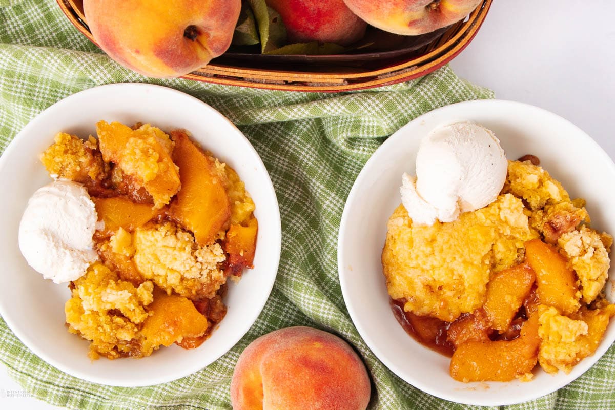Two bowls of peach cobbler topped with scoops of vanilla ice cream sit on a green checkered cloth, with fresh peaches and a basket of peaches in the background.