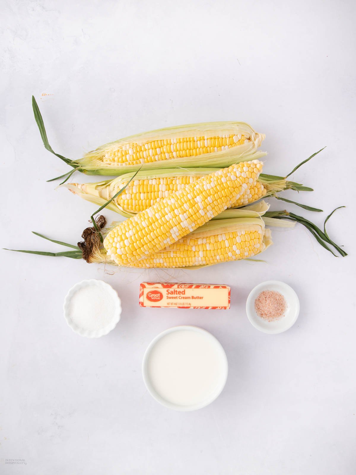 Four ears of fresh corn, a stick of salted butter, a small bowl of sugar, a small bowl of salt, and a bowl of cream are arranged on a white surface.