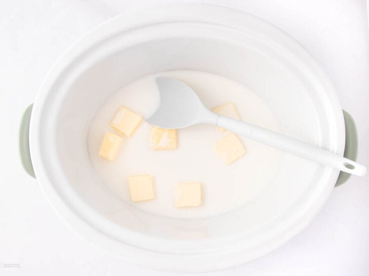 A white slow cooker with several cubes of butter and milk inside. A white spatula rests on the rim, ready for stirring.