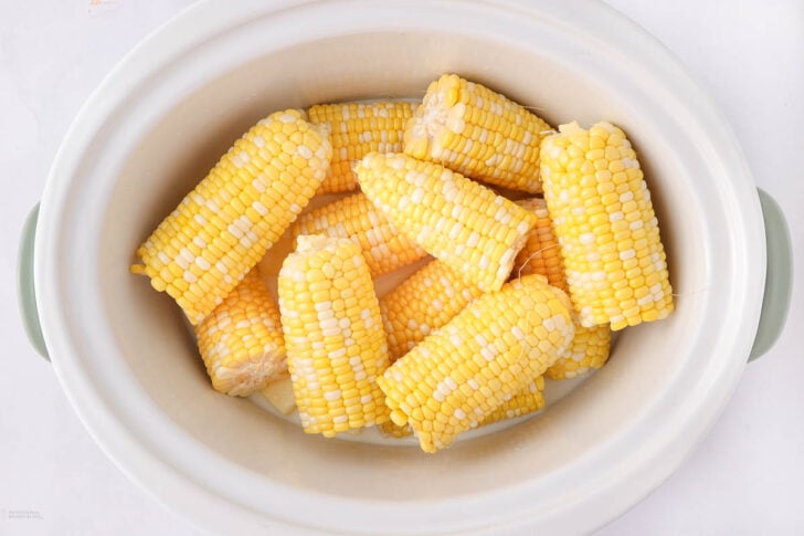 Several pieces of corn on the cob, cut into halves, are arranged inside a white slow cooker. The corn kernels are yellow and white, and the cooker is seen from above.