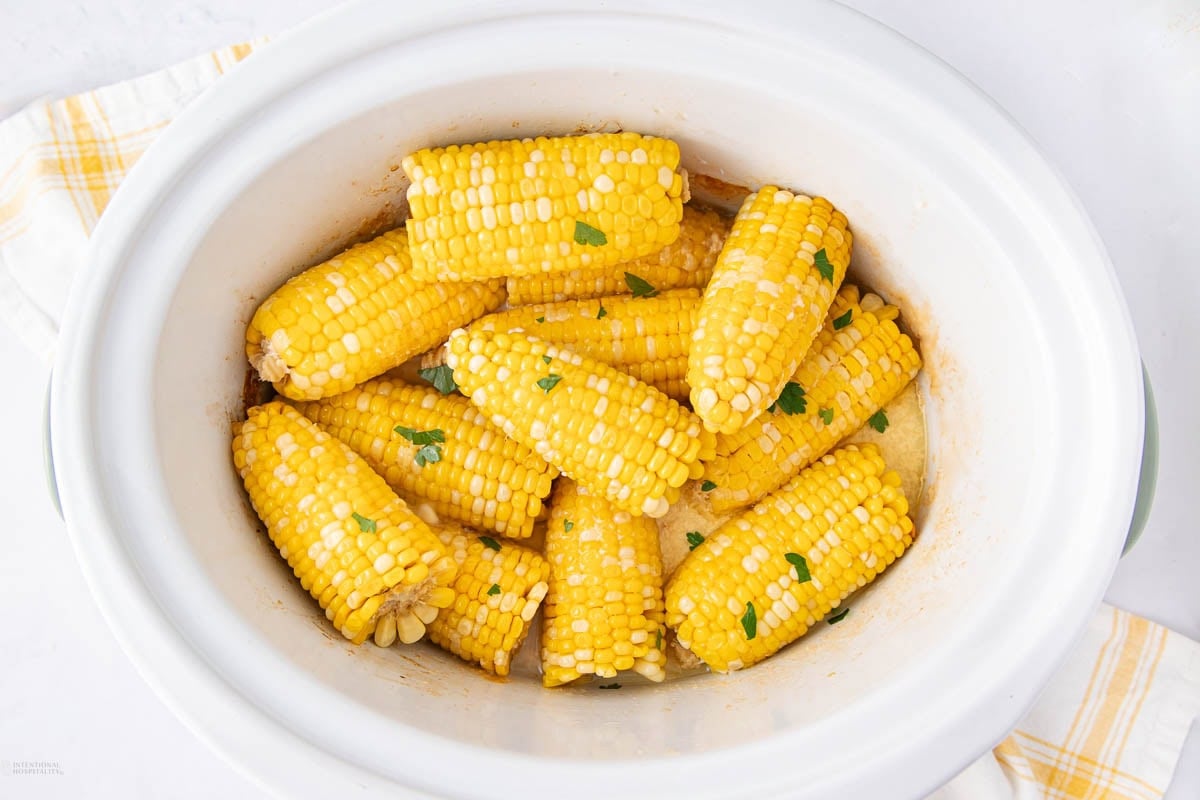 Corn on the cob pieces cooked in a white slow cooker, garnished with chopped herbs and placed on a white surface with a yellow plaid napkin nearby.