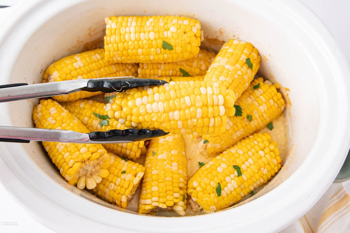 A slow cooker filled with corn on the cob halves garnished with herbs, with metal tongs holding one piece above the rest.