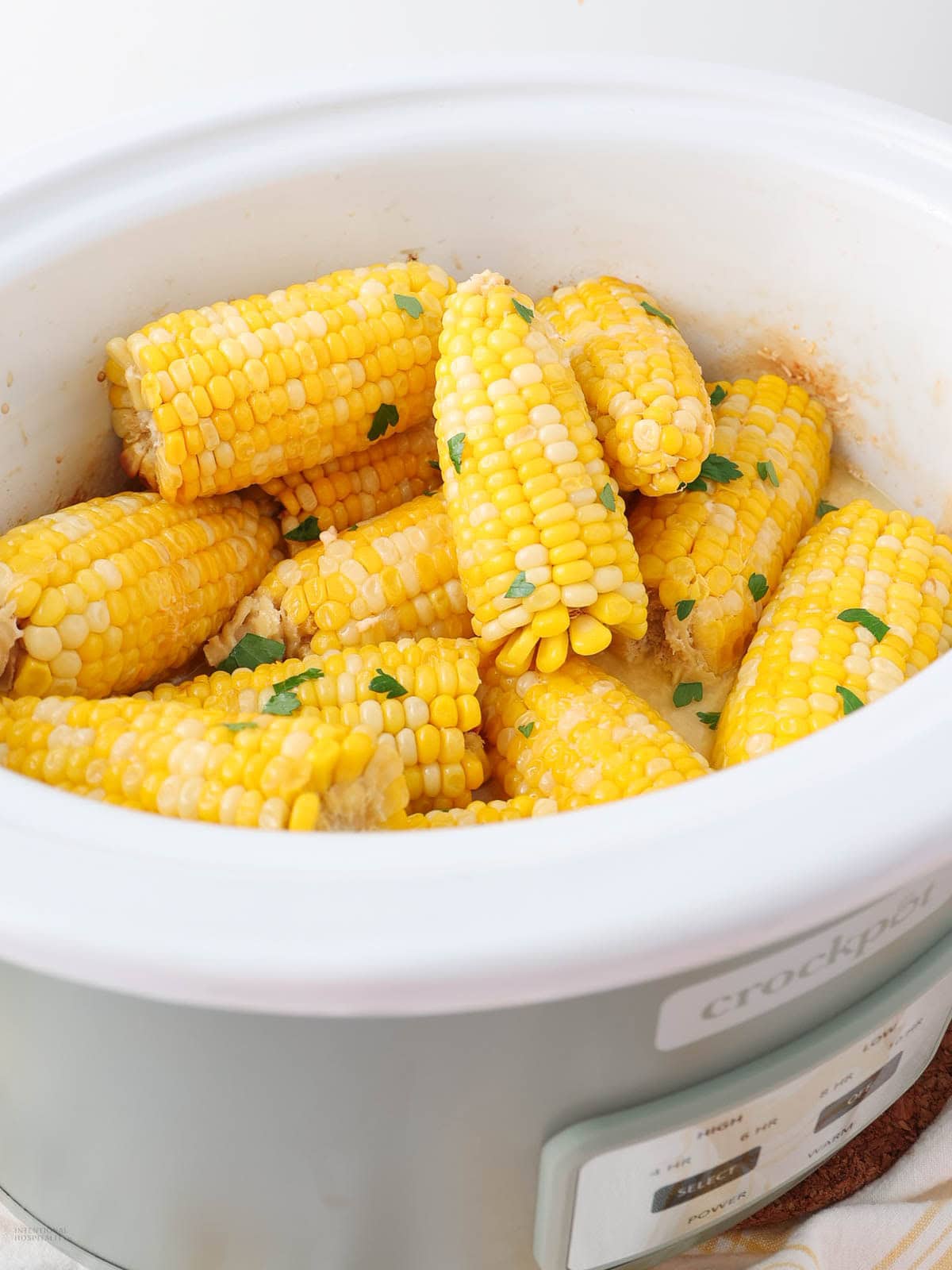 Corn on the cob pieces cooked in a white crockpot, garnished with chopped herbs.